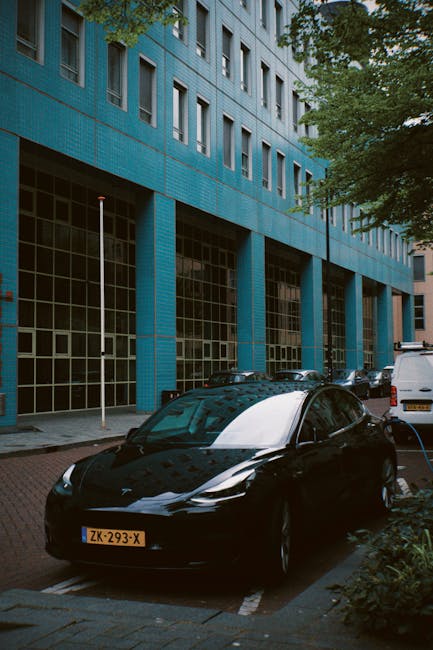 The image depicts the exterior of a modern blue residential or commercial building with large glass windows and vertical columns. The ground level features a parking area where a sleek black electric car with a shiny surface is parked, along with a white van and other vehicles. Surrounding the building are trees with green foliage, providing natural shading. The pavement appears clean and well-maintained, with clearly marked parking spaces. The scene is illuminated by natural daylight, highlighting the reflective surfaces of the vehicles and the building's glass, suggesting a tidy and orderly environment. This setting illustrates the importance of maintaining clean outdoor spaces and vehicle surfaces, consistent with the cleaning expertise provided by Cleaners Holland Park, especially relevant for buildings and parking areas on Holland Park terraces as mentioned in the page title.