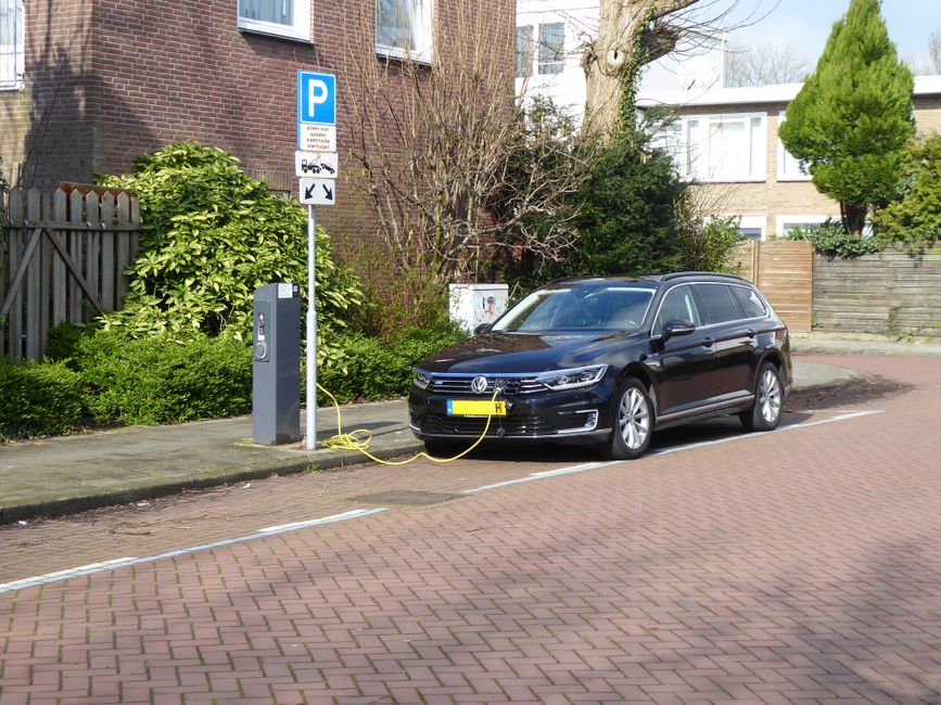 A black Volkswagen station wagon is parked in a designated electric vehicle charging spot on a paved residential street, connected to a public charging station with a yellow charging cable. The scene is well-lit, showing the car in front of a brick house with a wooden fence, surrounded by greenery including bushes and a large tree with bare branches. The surface of the road is clean and smooth, with white lines marking the parking area. The charging station features a digital display and control button, emphasizing the modern, environmentally friendly setting, consistent with hygienic and maintenance standards associated with professional cleaning services by Cleaners Holland Park.
