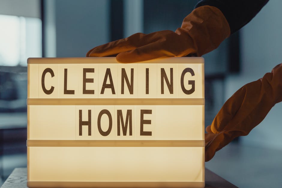 A person wearing orange gloves is holding a lightbox sign displaying the words 'CLEANING HOME' in bold, black letters. The sign is situated on a clean, smooth surface, with the background featuring blurred indoor elements and soft lighting. The image emphasizes surface cleaning and sanitisation, relevant to residential cleaning services, and is associated with Cleaners Holland Park, highlighting professional domestic cleaning efforts to maintain hygiene and shine in homes, free from hidden charges, as described in the page about avoiding hidden cleaning charges in Holland Park service quotes.