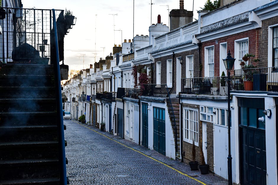 A row of terraced houses in Holland Park with brick and white-painted facades, featuring large windows and small balconies with black wrought iron railings. The street is paved with cobblestones and gently slopes upward, illuminated by soft natural light from the setting sun. Visible elements include potted plants on balconies, street lamps, and external staircases. The overall scene reflects a clean and well-maintained residential area, with no visible dirt or debris, emphasizing the importance of surface cleaning and maintenance. This image showcases the charming appearance of traditional UK townhouses, aligning with the concept of domestic cleaning as part of a comprehensive end of tenancy cleaning checklist, provided by Cleaners Holland Park.