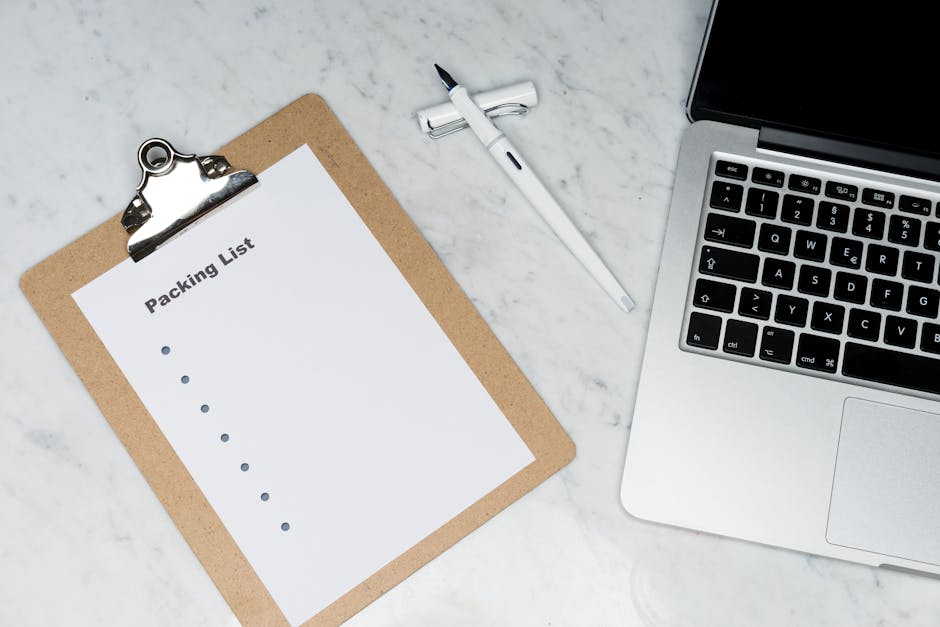 A white sheet titled 'Packing List' clipped to a light brown clipboard, placed on a marble countertop. To the right, there is a white marker or pen lying horizontally, and an open silver laptop with a visible keyboard and a black screen. The surface appears clean, well-lit, and arranged systematically, indicating an organized setup for preparing an end of tenancy cleaning checklist. This scene is part of a domestic cleaning or preparation process, with the focus on organization and hygiene, and relates to the services offered by Cleaners Holland Park for comprehensive cleaning and sanitisation tasks. The workspace is minimalistic, emphasizing tidiness and readiness for detailed cleaning planning as per the page titled 'Campden Hill end of tenancy cleaning checklist, Holland Park' available at cleaners-hollandpark.com.