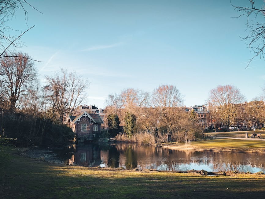 The image shows a tranquil outdoor scene in a park during autumn, featuring a narrow, calm river with reflective surface. Surrounding the river are deciduous trees with yellow and orange foliage, some