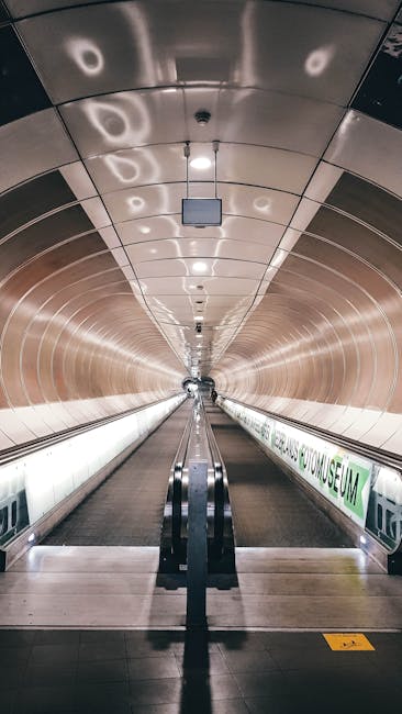A long, enclosed walkway leading to Holland Park station, with curved, metallic walls and ceiling panels that reflect light, creating a shiny, polished appearance. The floor features a dark, textured surface with a central moving walkway flanked by glass barriers and handrails. Overhead lighting fixtures are evenly spaced along the ceiling, illuminating the tunnel with bright, white light. The space appears clean, hygienic, and well-maintained, consistent with professional deep cleaning standards provided by Cleaners Holland Park for efficient surface cleaning and sanitisation of transit environments.