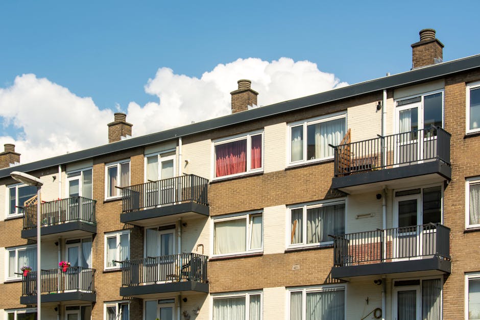 Exterior view of a three-story residential apartment building in Holland Park, featuring a brick façade with multiple floor-to-ceiling windows, some with white curtains. Small balconies with metal rai